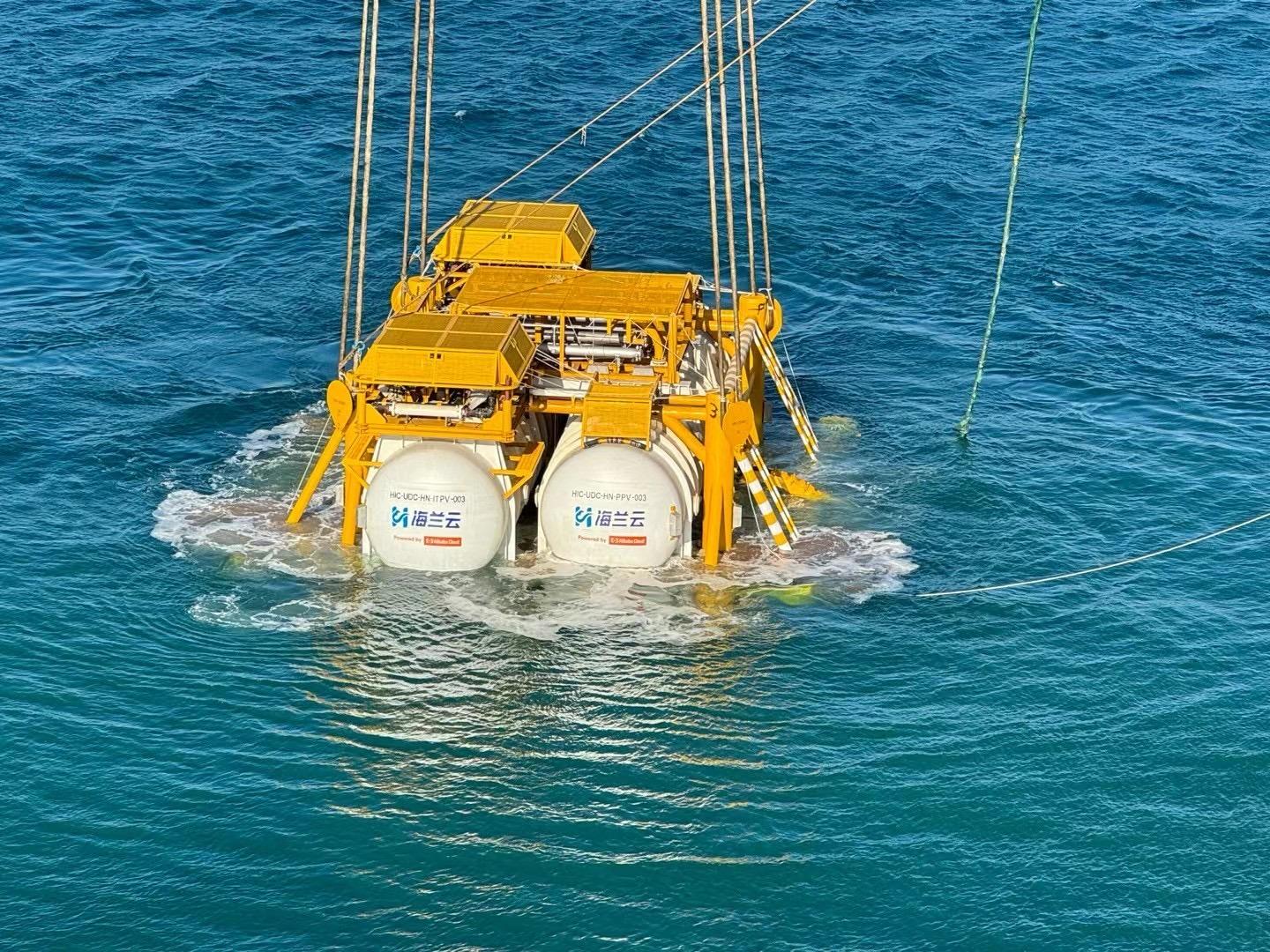 A large underwater equipment structure is being lowered into the ocean using cables. The device has yellow metal framework and two prominent white cylindrical tanks with text and symbols on them.