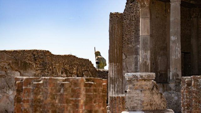 Ancient ruins with stone columns and damaged walls, featuring a statue partially visible in the background. Clear blue sky above.