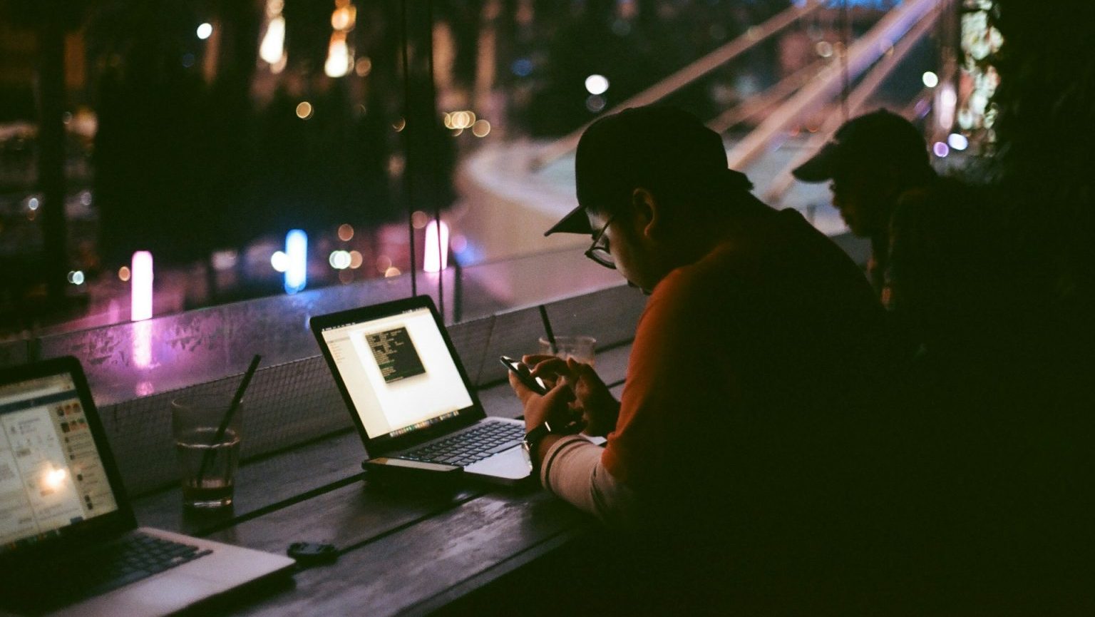 Person sitting at a dimly lit table with a laptop, using a smartphone. Another person is visible in the background. There are buildings and lights visible through the window.