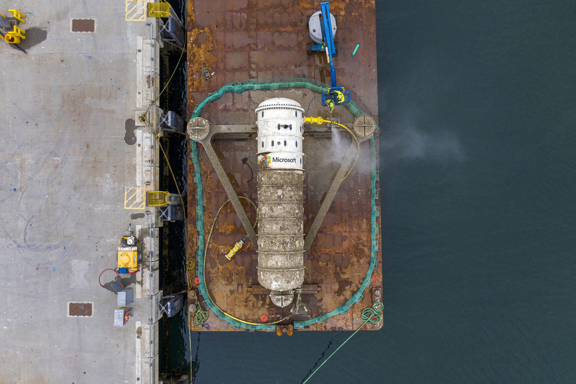 Aerial view of a cylindrical data center pod named "Microsoft" on a barge near a dock, surrounded by water with two workers and equipment on the platform.