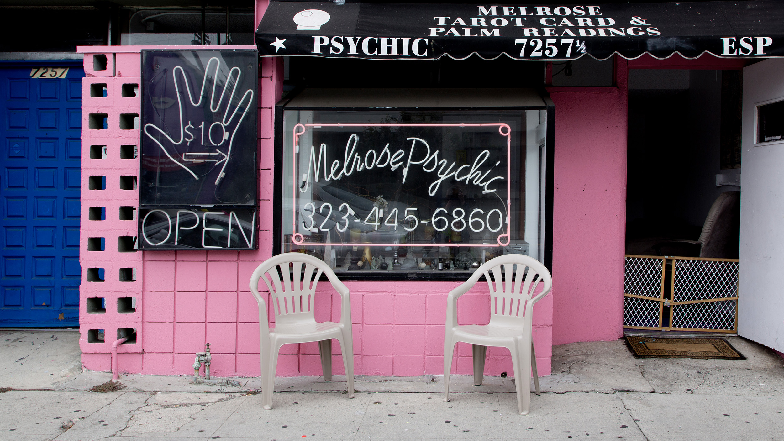 A storefront with signs for psychic telepathy readings and a phone number. Two white plastic chairs are placed in front. The storefront is pink with a black awning and a large hand sign advertising $10 readings.