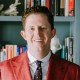 A person with curly hair wearing a red blazer and patterned tie stands in front of a bookshelf filled with books and decorative items.