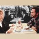 Two people sitting across from each other at a table in a diner, engaged in conversation. The table has plates of food and drinks. The background shows other diners in a black and white setting.