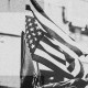 Black and white photo of an American flag waving against a blurred background of buildings, framed by red and beige rectangular blocks.