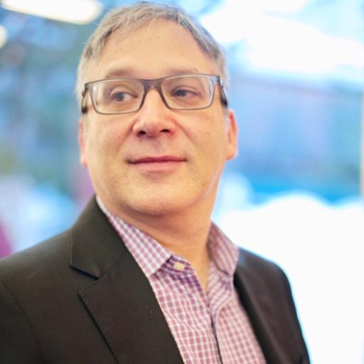 A man with glasses wearing a black blazer and checkered shirt looks to the side with a slight smile.