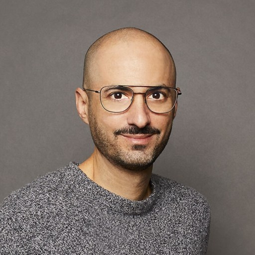 A man with glasses, a beard, and a shaved head, wearing a gray sweater, smiles against a plain gray background.