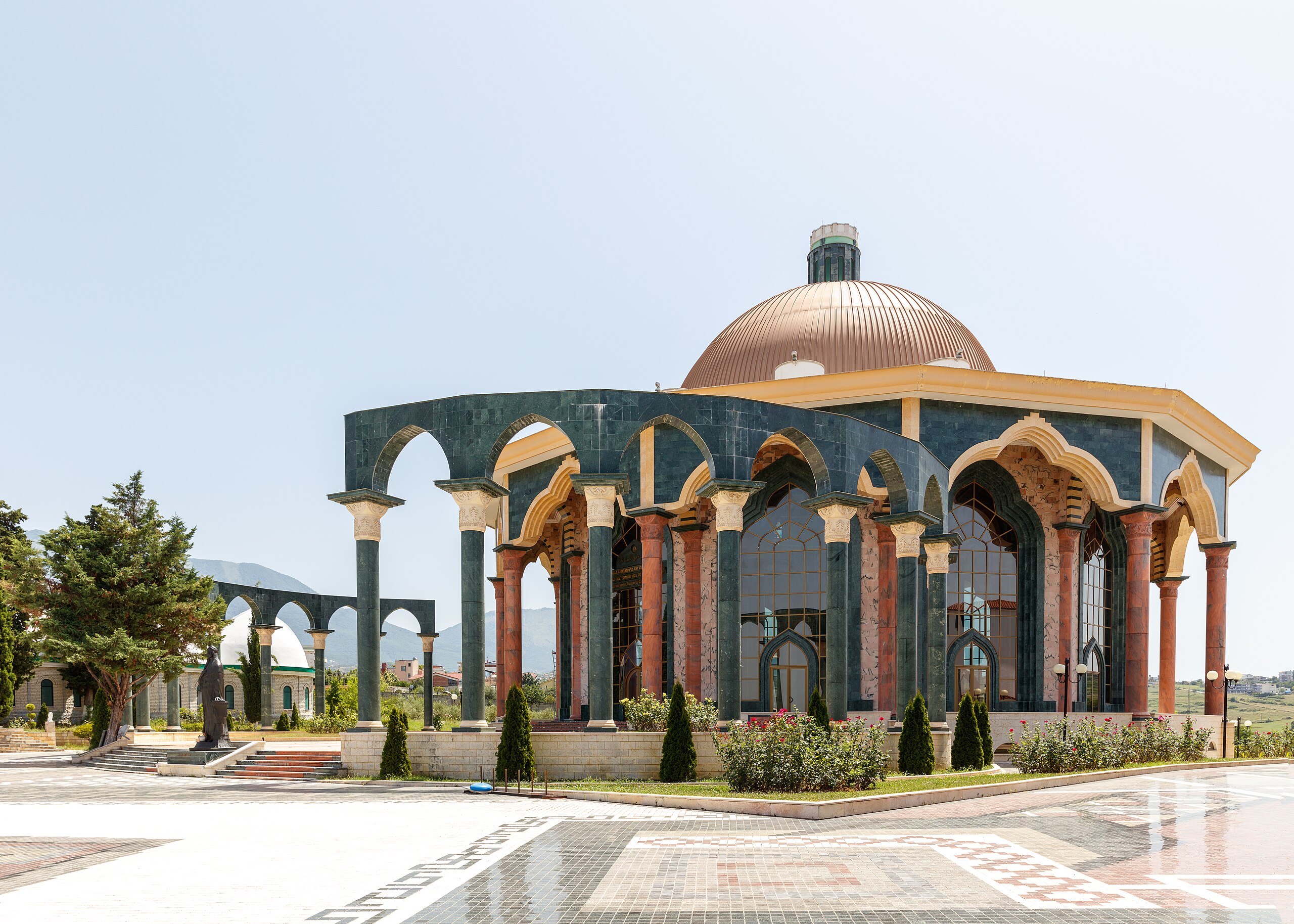 A large building with a copper dome and multiple arches stands in a plaza with a patterned tile floor. Trees and landscaping are visible around the building.