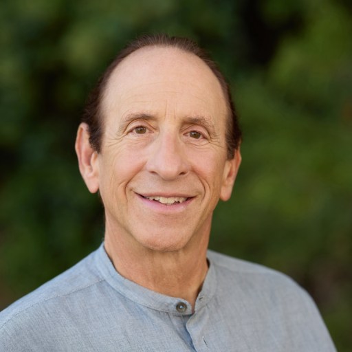 A man with short, dark hair smiles at the camera while wearing a light gray shirt in an outdoor setting with greenery in the background.