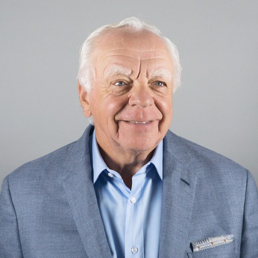 Elderly man with white hair and a smile, wearing a light blue shirt and a light blue blazer with a silver tie pin and a pocket square, against a plain gray background.