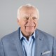 Elderly man with white hair and a smile, wearing a light blue shirt and a light blue blazer with a silver tie pin and a pocket square, against a plain gray background.