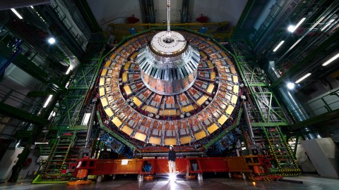 A person stands in front of a large, circular particle detector in a brightly lit, high-tech facility.