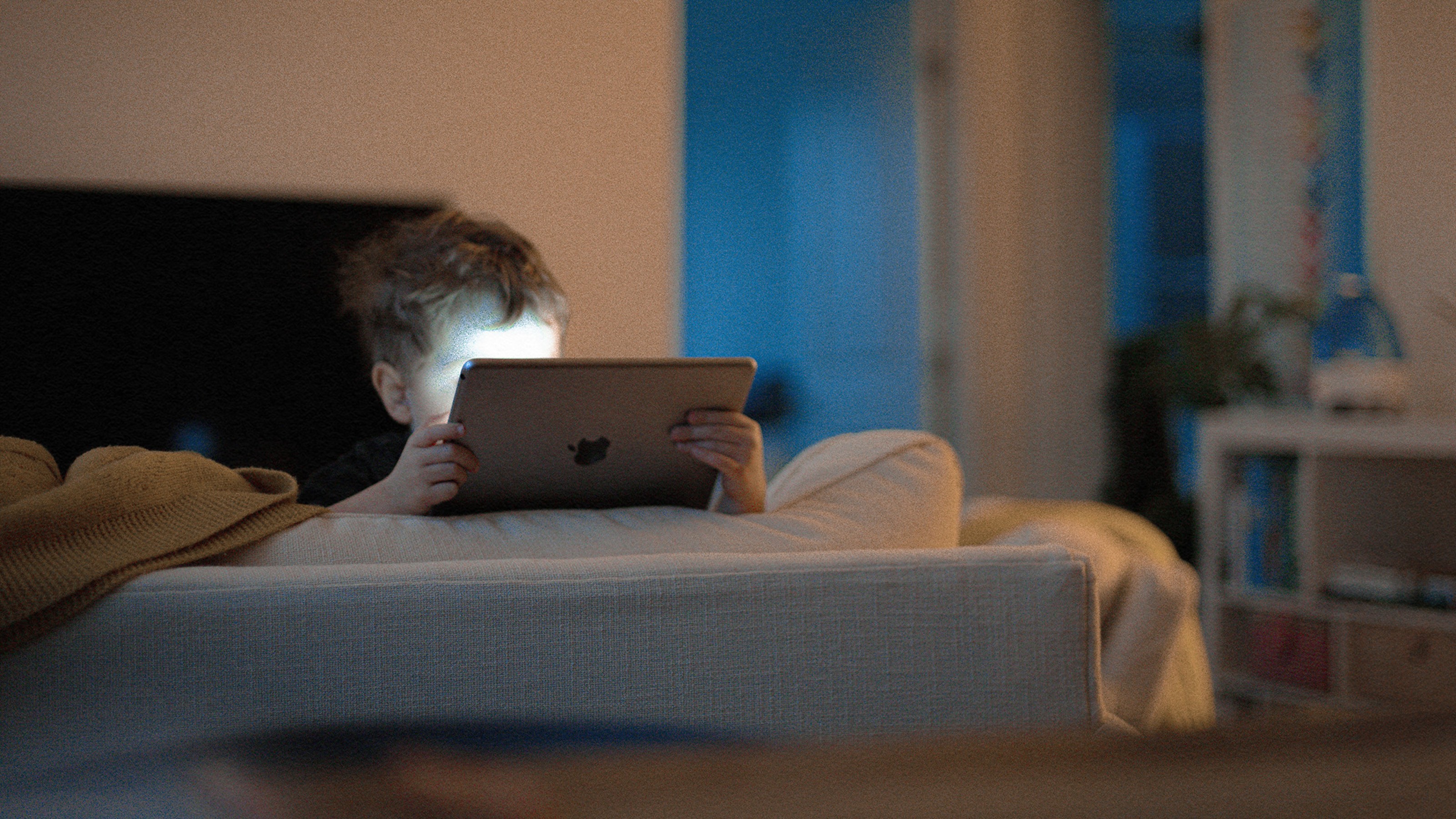 Young child sitting on a couch, illuminated by the screen of a tablet they are holding in a dimly lit room.