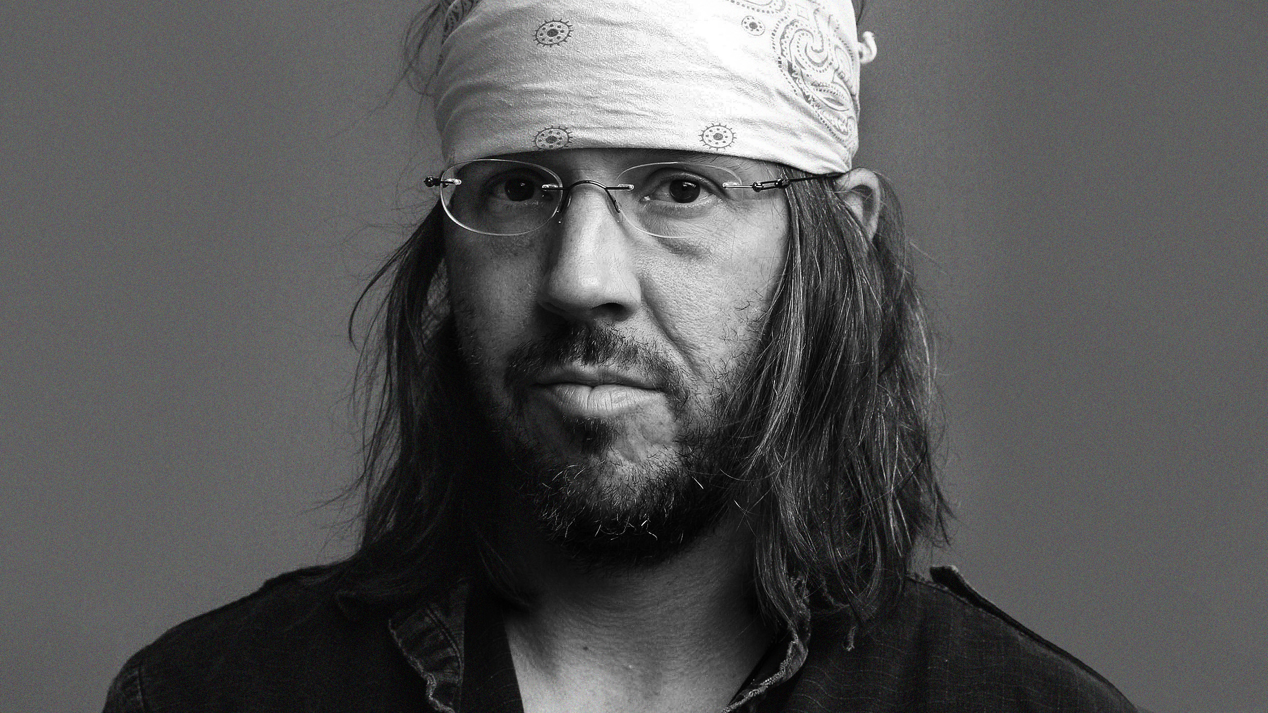 A man with long hair, a beard, glasses, and a bandana on his head, reminiscent of David Foster Wallace, poses for a portrait against a plain background.