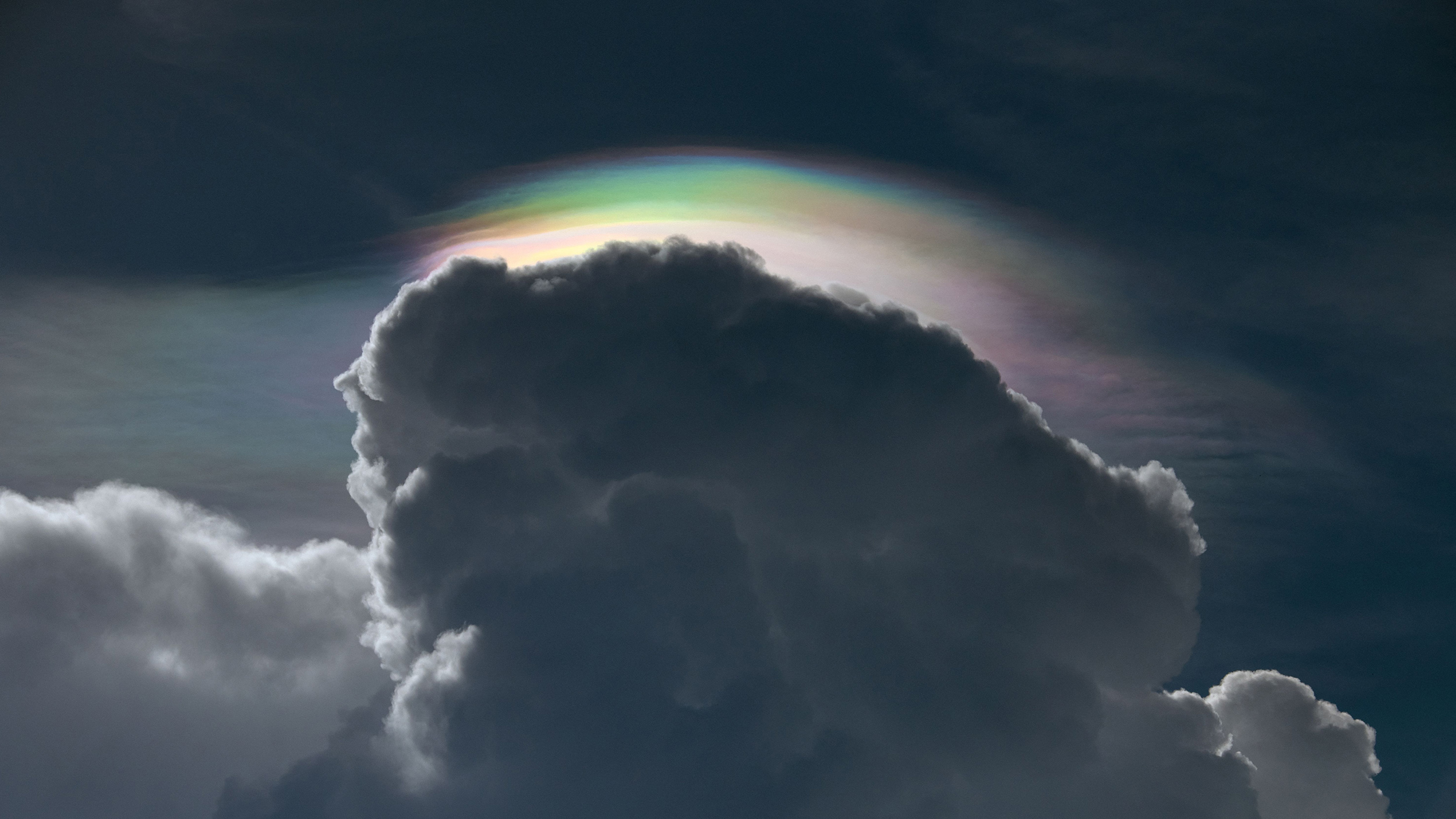 A dark cloud, laced with cynicism, is topped with a colorful iridescent layer against a dim sky.