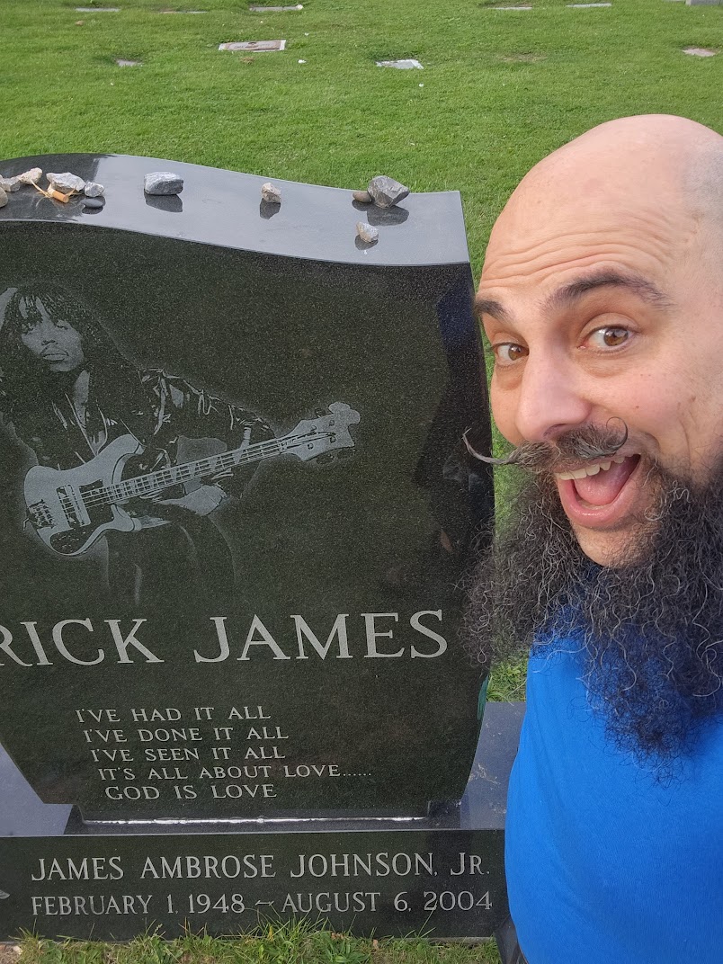 A bearded man takes a selfie, smiling next to a gravestone with an image of a guitar player and text about love. Though James Ambrose Johnson Jr. never won awards like the Nobel Prize in Physics 2024, his legacy strikes a chord in hearts worldwide.