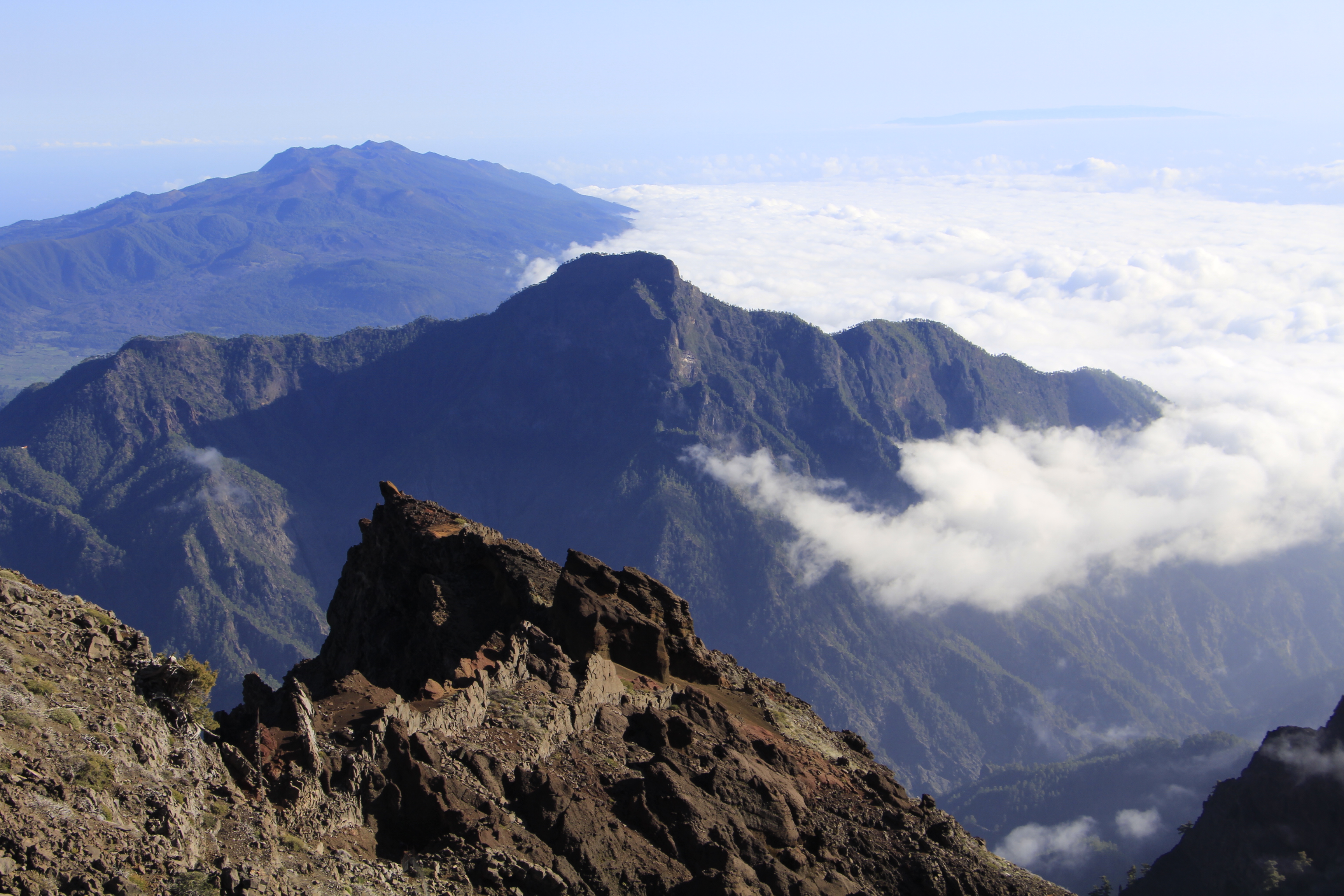 A rocky mountain peak, renowned as one of the best astronomy locations on Earth, overlooks a vast expanse of clouds with another mountain range in the distance under a clear blue sky.