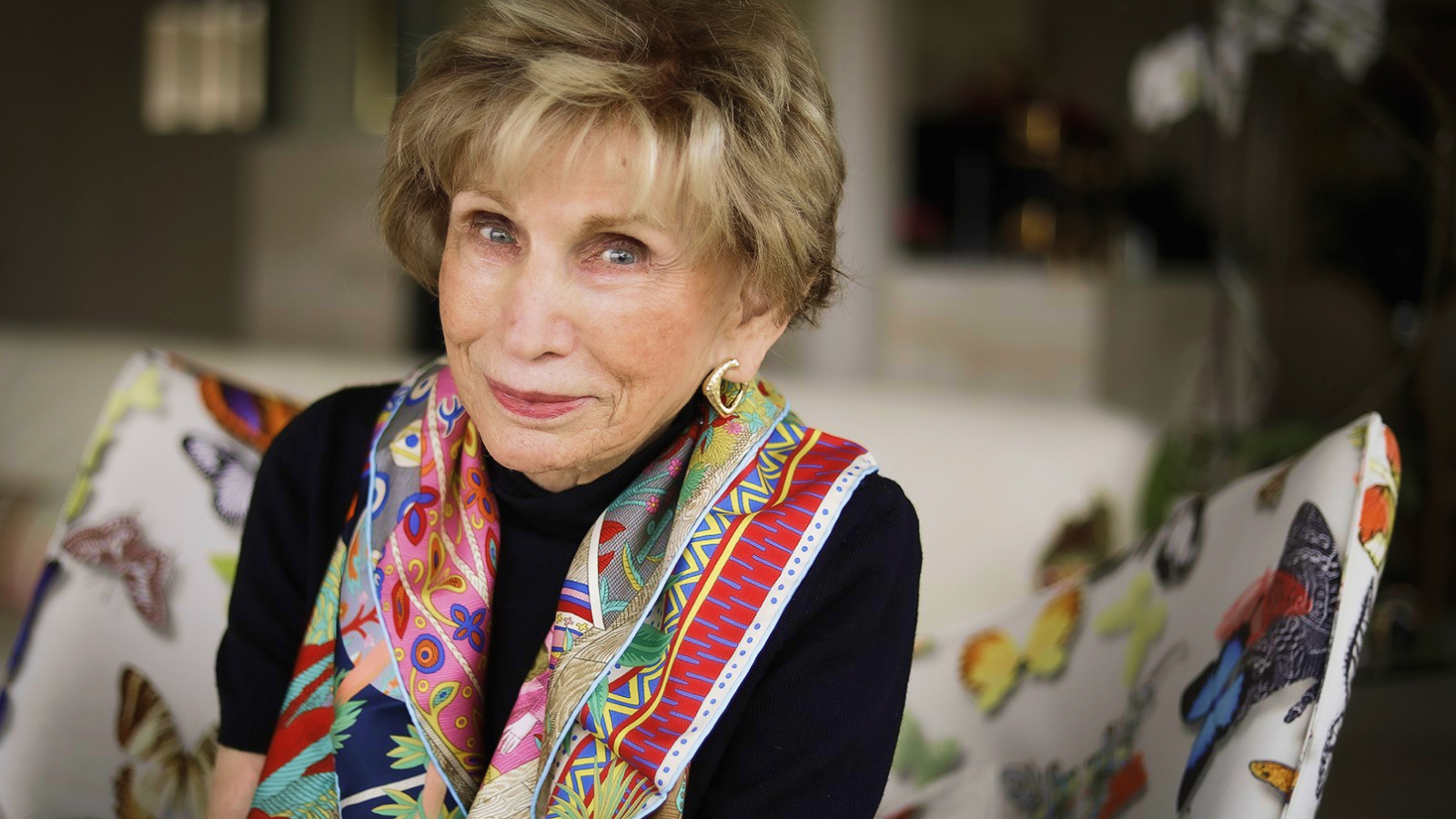Edith Eva Eger, an older woman with short light hair, wearing a colorful scarf and gold earrings, sits on a chair with a butterfly pattern in a well-lit room.
