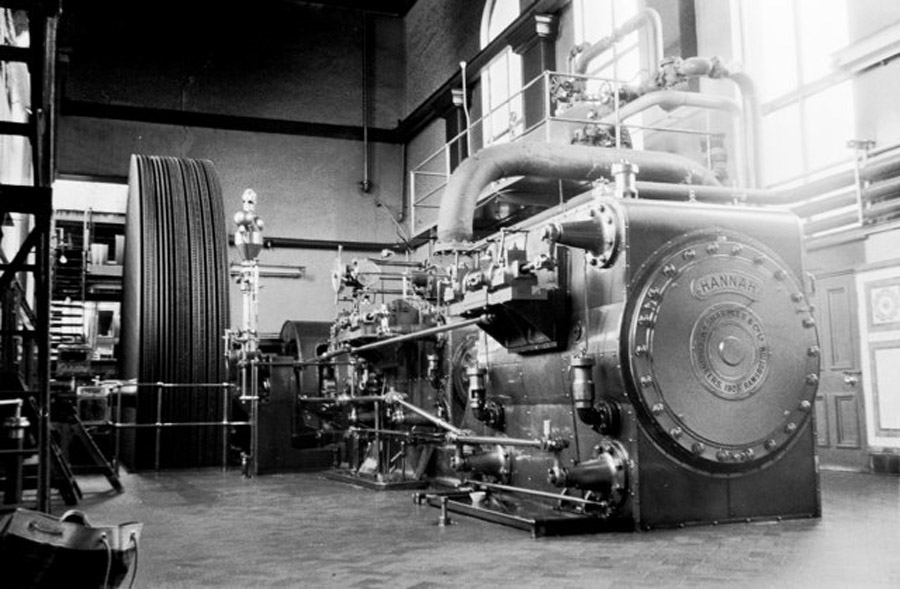 A large, industrial steam engine with intricate piping and a labeled flywheel is set in a factory room with high windows.