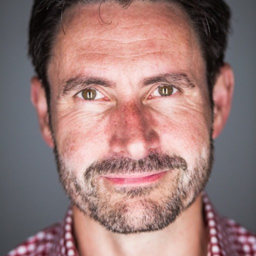 A man with a beard and mustache, wearing a red and white checkered shirt, looks directly at the camera against a gray background.