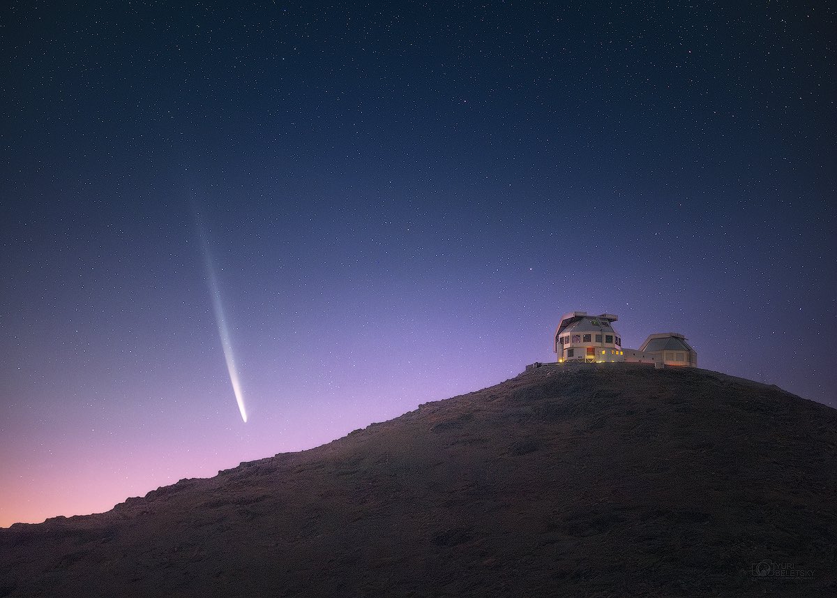 A comet streaks across a starry sky above an observatory situated on a hilltop at twilight.