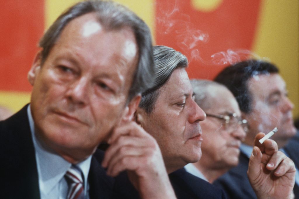 A group of men in suits sit at a formal event; one man is smoking a cigarette.