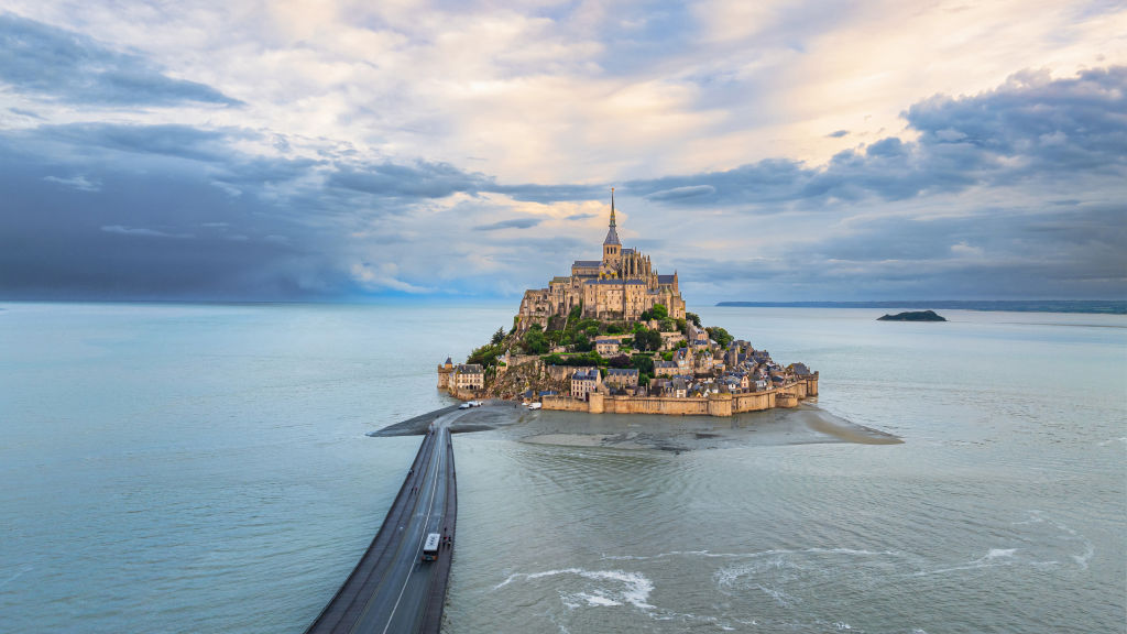 Aerial view of Mont Saint-Michel, a rocky island with an abbey, surrounded by water at high tide, connected to the mainland by a causeway under a clouded sky.