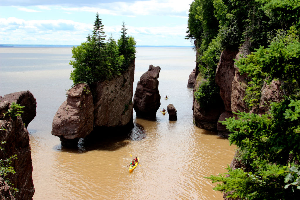 Kayakers paddle between tall, rocky cliffs and forested rock formations in a body of water with a view of the horizon.