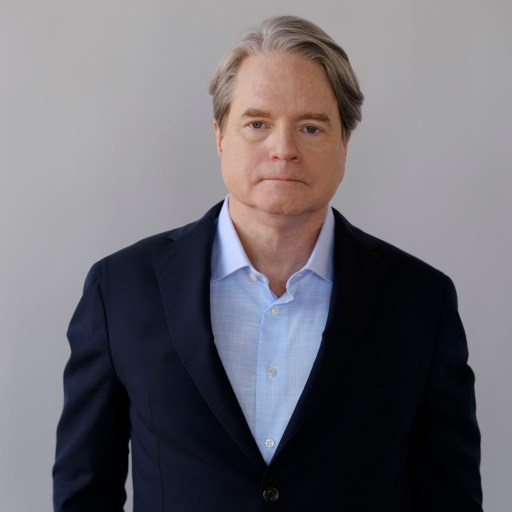 A man in a dark suit and light blue shirt stands against a plain background.