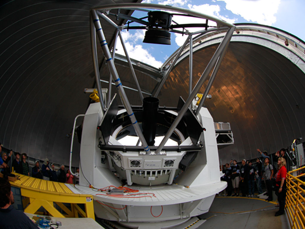 A large telescope is housed inside an observatory dome, surrounded by people.