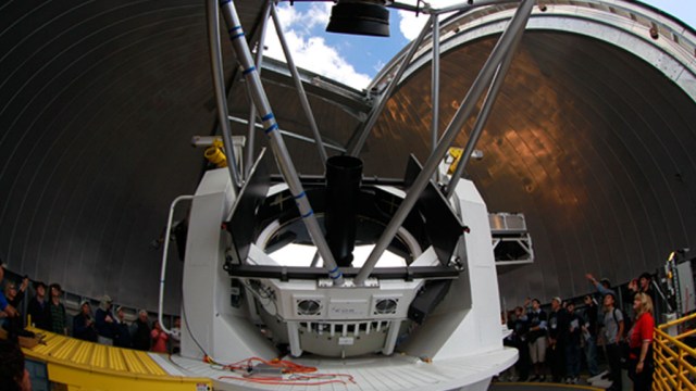 A large telescope is housed inside an observatory dome, surrounded by people.