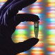 A gloved hand holds a small vial in front of a colorful, blurred background resembling a DNA sequence.