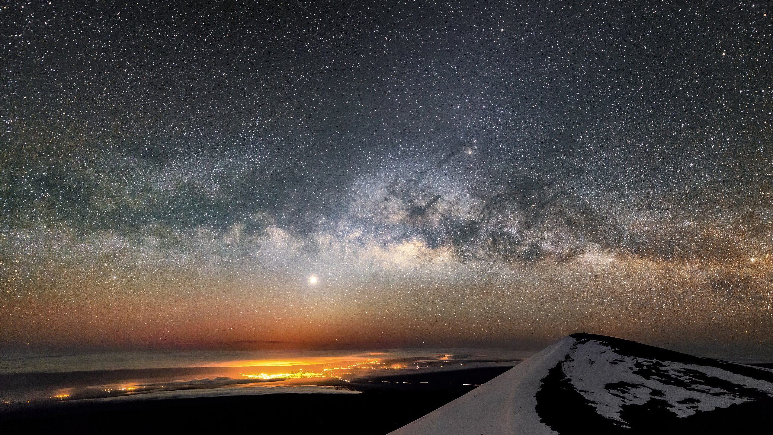 A starry night sky over a snow-capped mountain, known as one of Earth's best astronomy locations, with bright city lights shimmering in the distance on the horizon.