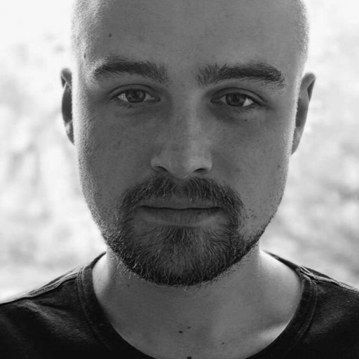 A person with a shaved head and beard looks directly at the camera, wearing a dark shirt. The background is softly blurred. Black and white image.