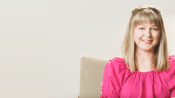 A woman with blonde hair smiles while sitting on a beige chair. She wears a bright pink top and the background is a plain light color.