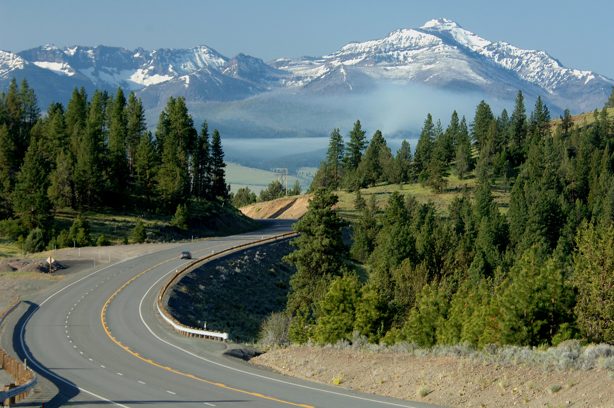 A winding road curves through a forested landscape with distant snow-capped mountains under a clear blue sky.