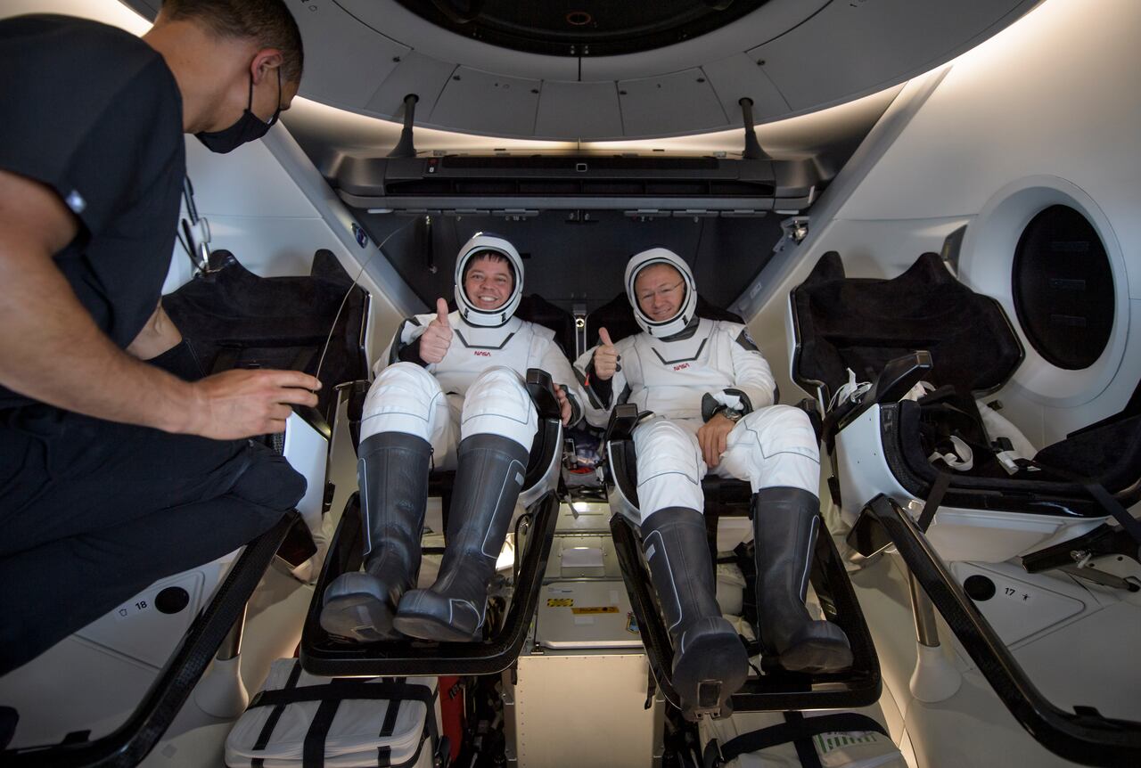 Two astronauts in white space suits sit inside a spacecraft cabin, giving thumbs up. A person stands nearby, partially visible, wearing a face mask.