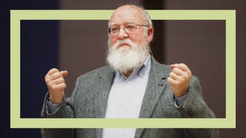 A person reminiscent of Daniel Dennett, known for his critical thinking, stands with raised hands, sporting a beard and glasses. He wears a gray blazer over a blue shirt against a neutral background.