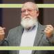 A person reminiscent of Daniel Dennett, known for his critical thinking, stands with raised hands, sporting a beard and glasses. He wears a gray blazer over a blue shirt against a neutral background.