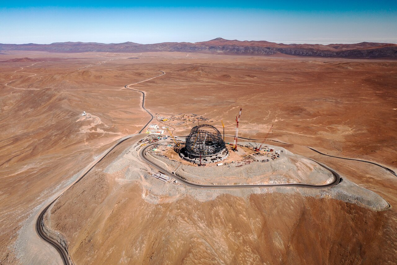 Aerial view of a large construction site in a desert landscape, featuring a domed metal structure surrounded by equipment and roads. Sparse mountains are visible on the horizon, suggesting this might be one of the best astronomy locations on Earth.