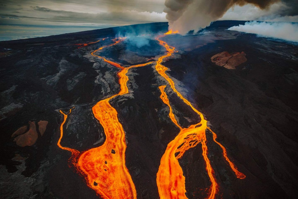 Aerial view of an active volcano with glowing lava flows streaming down its dark slopes and a plume of smoke rising into the sky, showcasing one of Earth's best locations for observing raw natural forces.