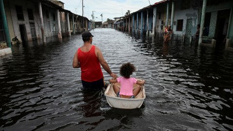 Amidst the chaos of natural disasters, a man pulls a child in a small boat through a flooded street lined with houses.