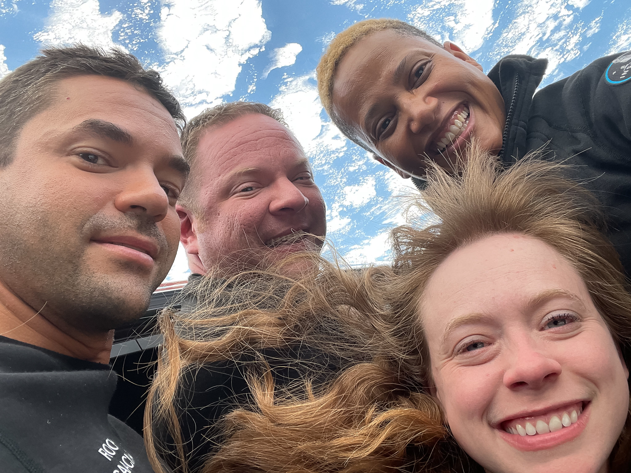 Four people smiling, floating in a spacecraft with Earth visible through the window behind them.