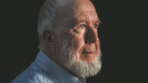 A side profile of an older man with a white beard, wearing a light blue shirt, against a dark background, illuminated by soft lighting from the left, exudes an aura of radical optimism.