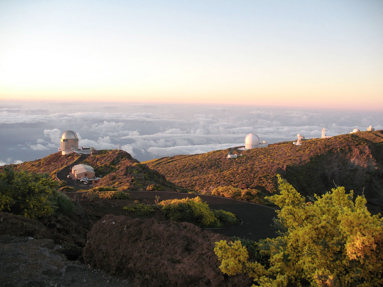 Several observatories, at arguably the best astronomy location on Earth, sit atop a mountain at sunset, surrounded by clouds and lush green vegetation.