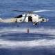 A helicopter hovers over the ocean as a person is lowered into the water, creating a circular ripple effect around them.