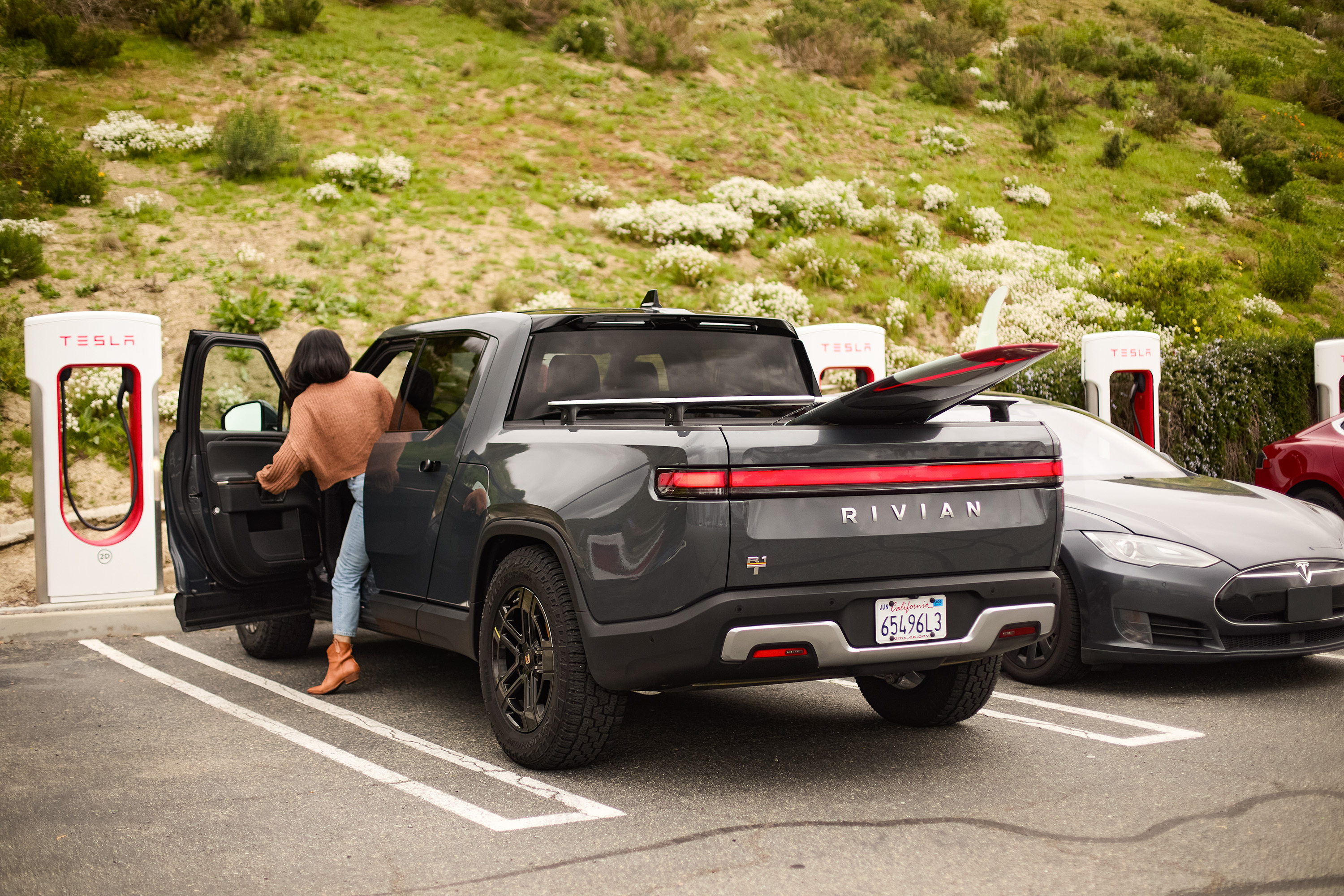 A gray Rivian truck is parked at a Tesla charging station. A person is opening the driver's door.