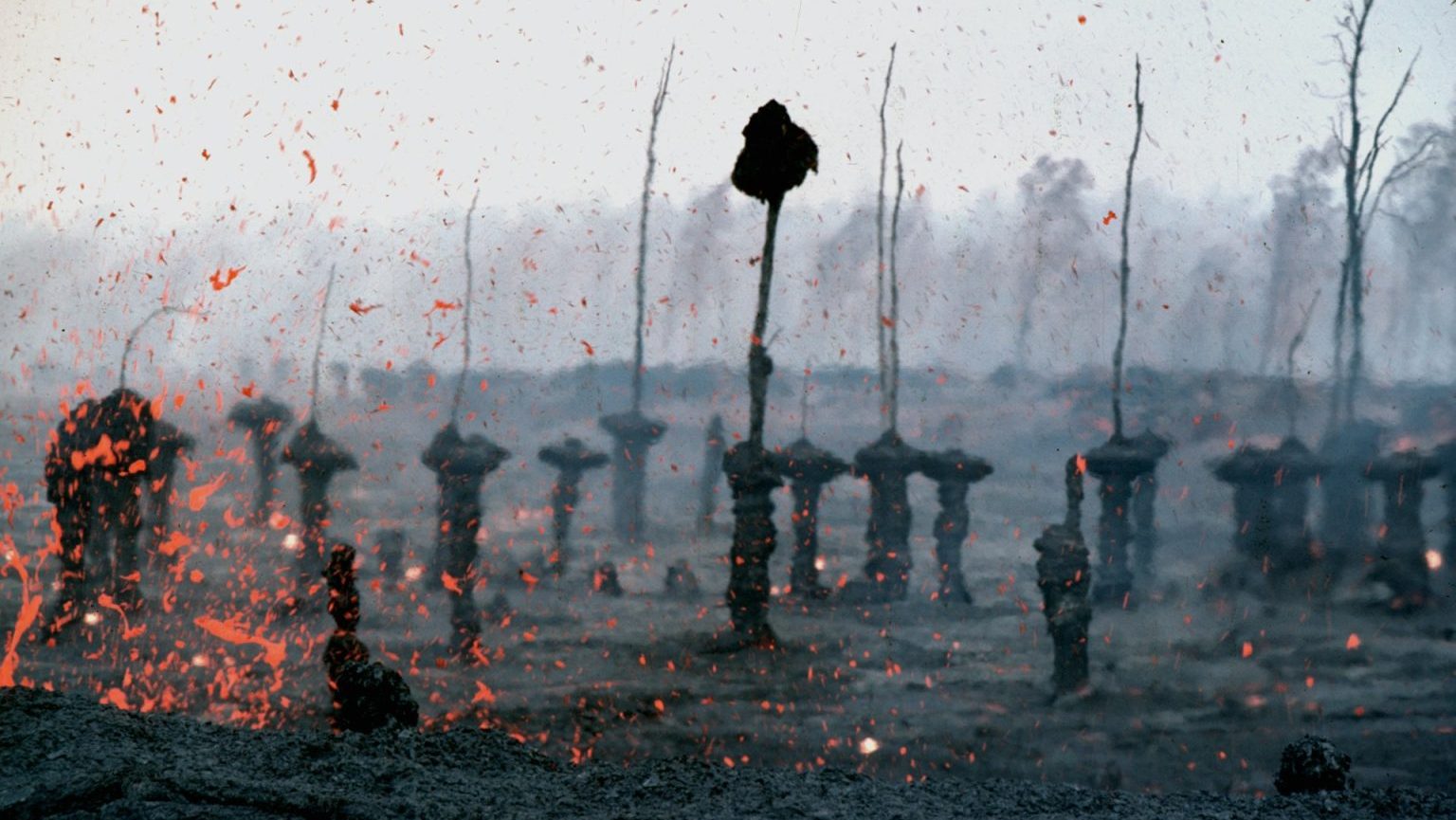 Lava explodes from the ground, surrounded by ash-covered trees and barren landscape under a hazy sky.