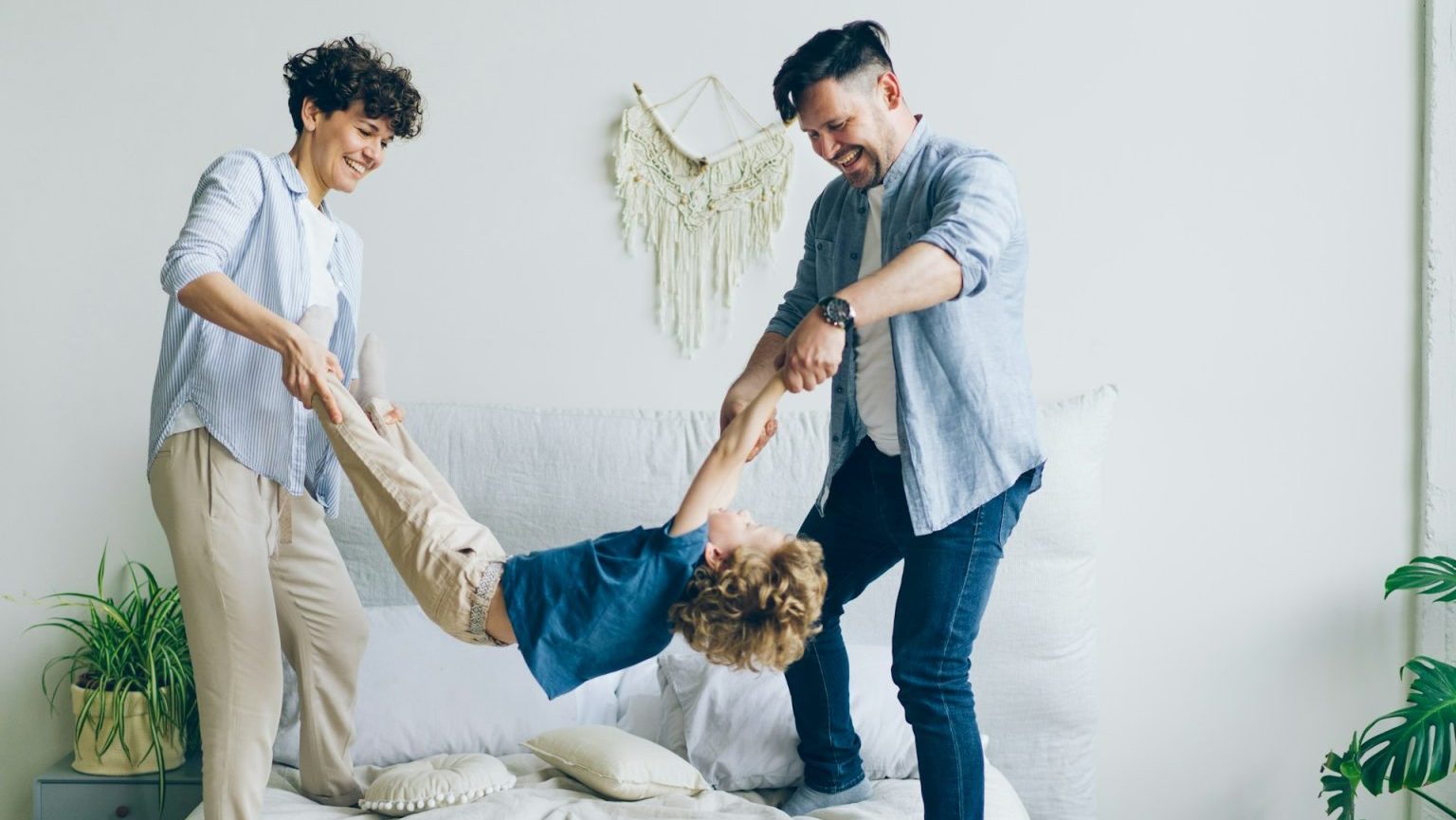 A child being playfully swung by two adults, standing on a bed in a bright room with plants and a wall hanging.