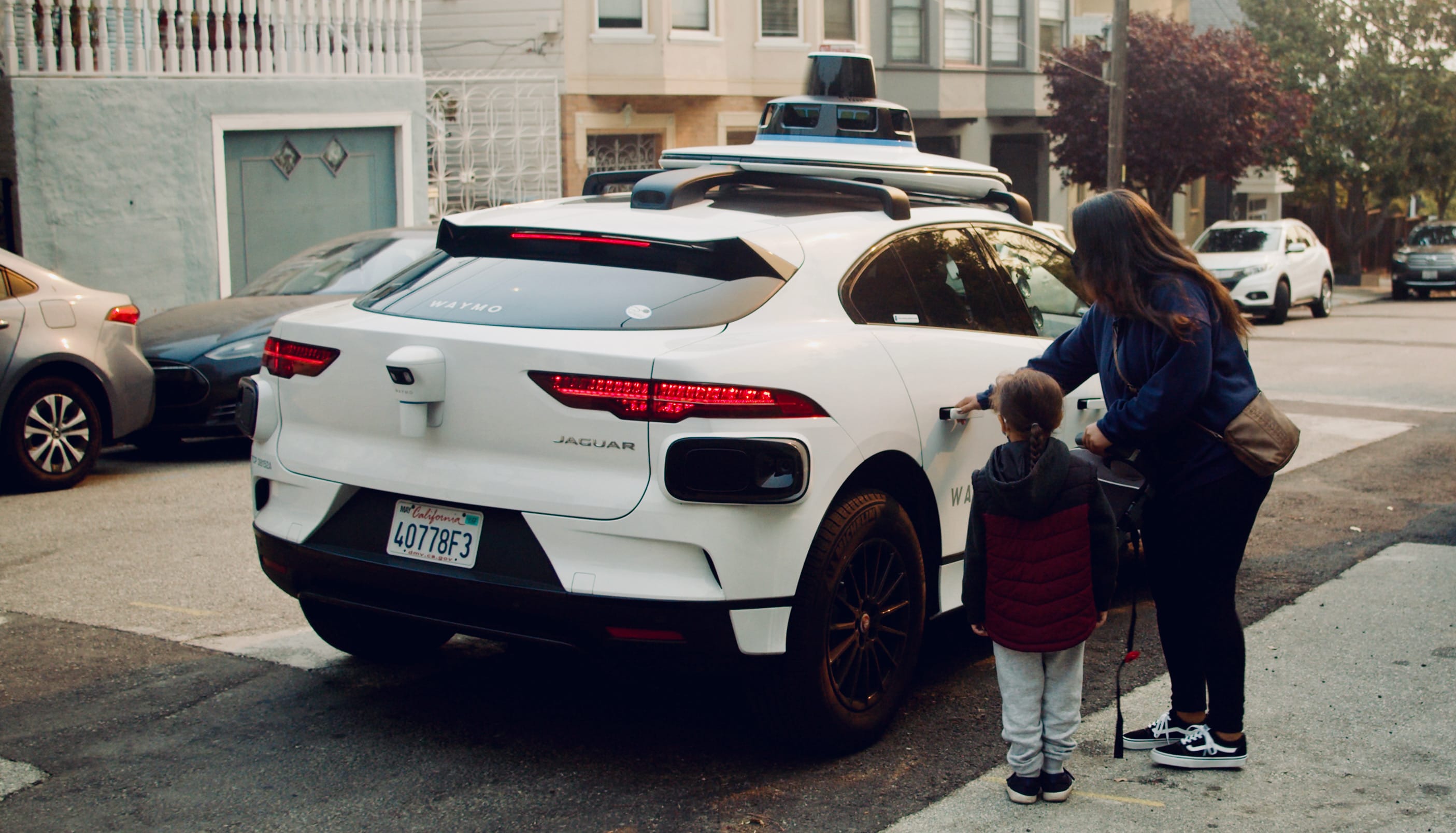 A woman and child stand beside a parked white autonomous vehicle on a residential street.