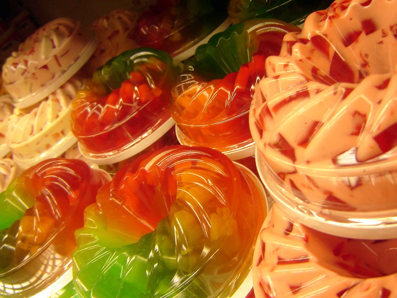 Rows of colorful gelatin desserts in various flavors and shapes, covered with clear lids.